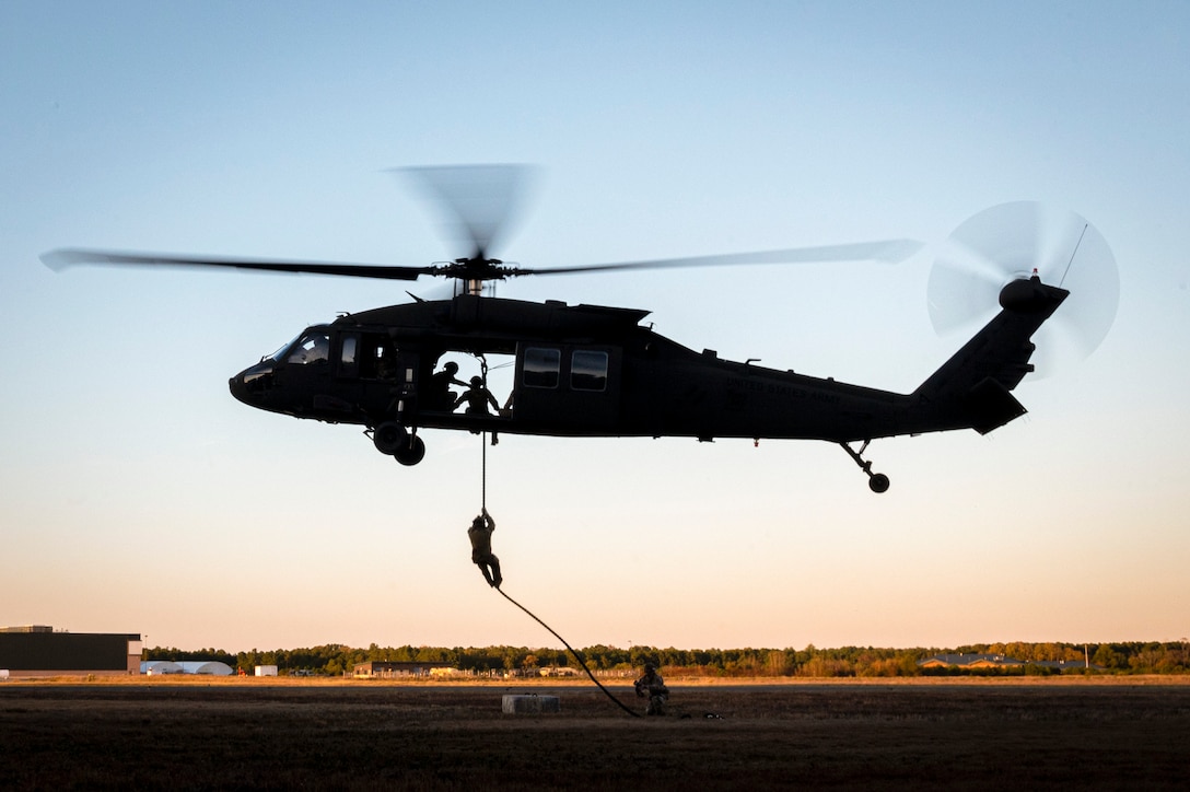 An Air National Guardsman fast-ropes from a helicopter in a field-like area as other service members watch.