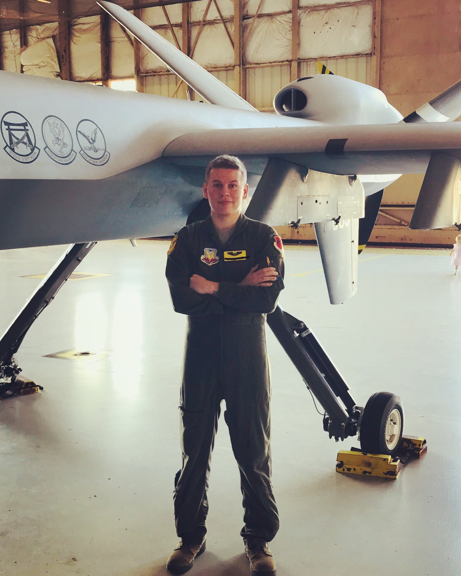 Airman 1st Class Jared, 482nd Attack Squadron, poses for a photo with his arms crossed while in front of an MQ-9 Reaper in a maintenance hanger.