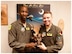 Airman 1st Class Jared, 482nd Attack Squadron, and Lt. Col. Andre, 482nd ATKS, hold a trophy together as they pose for a photo in front of a blue MQ-9 poster.