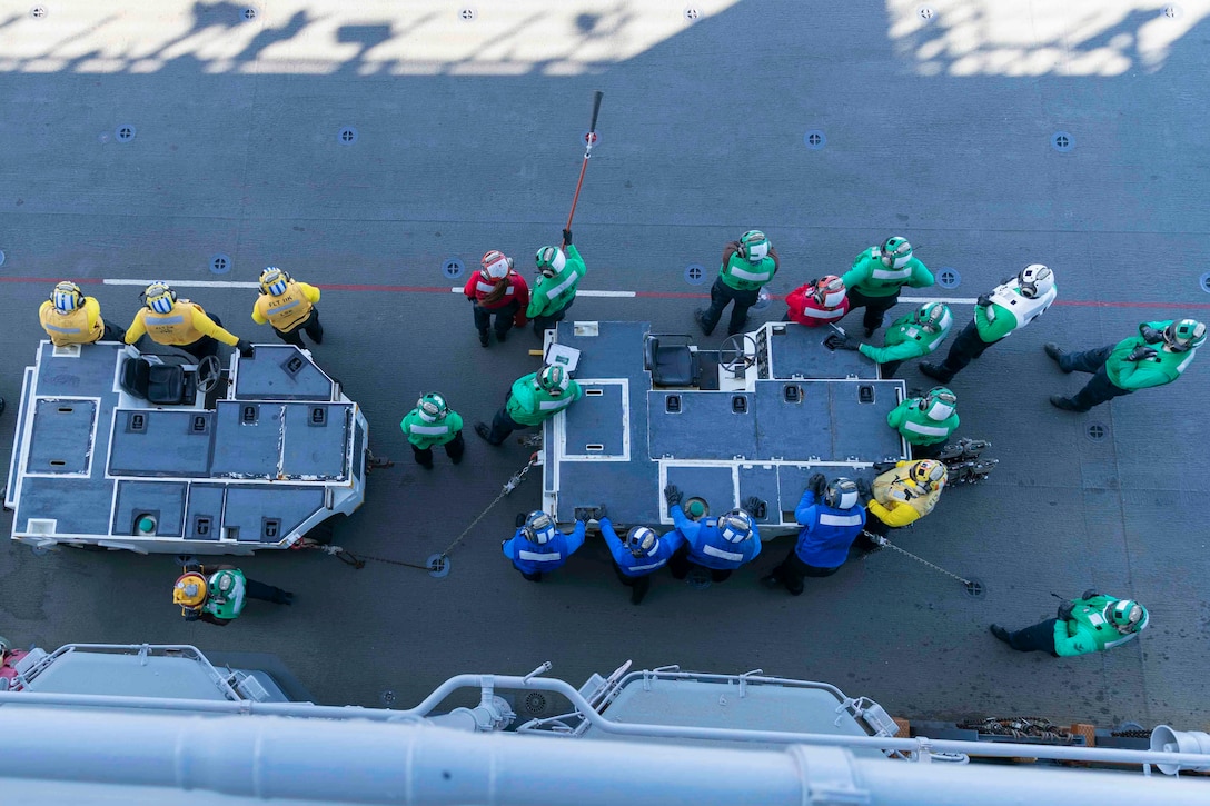Sailors wearing, seen from above, stand on ship's flight deck, wearing either yellow, red, blue or green shirts.