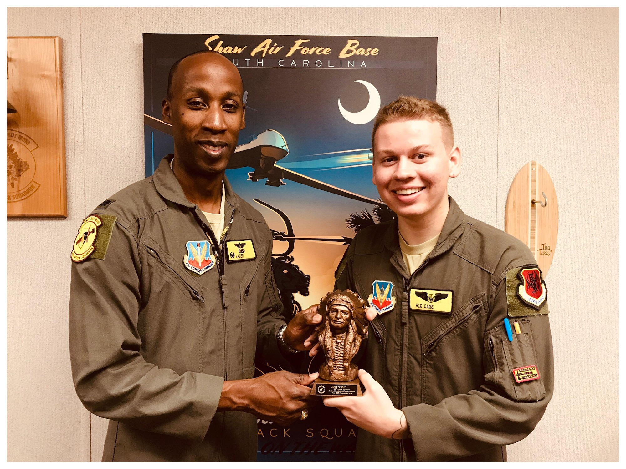 Airman 1st Class Jared, 482nd Attack Squadron, and Lt. Col. Andre, 482nd ATKS, hold a trophy together as they pose for a photo in front of a blue MQ-9 poster.