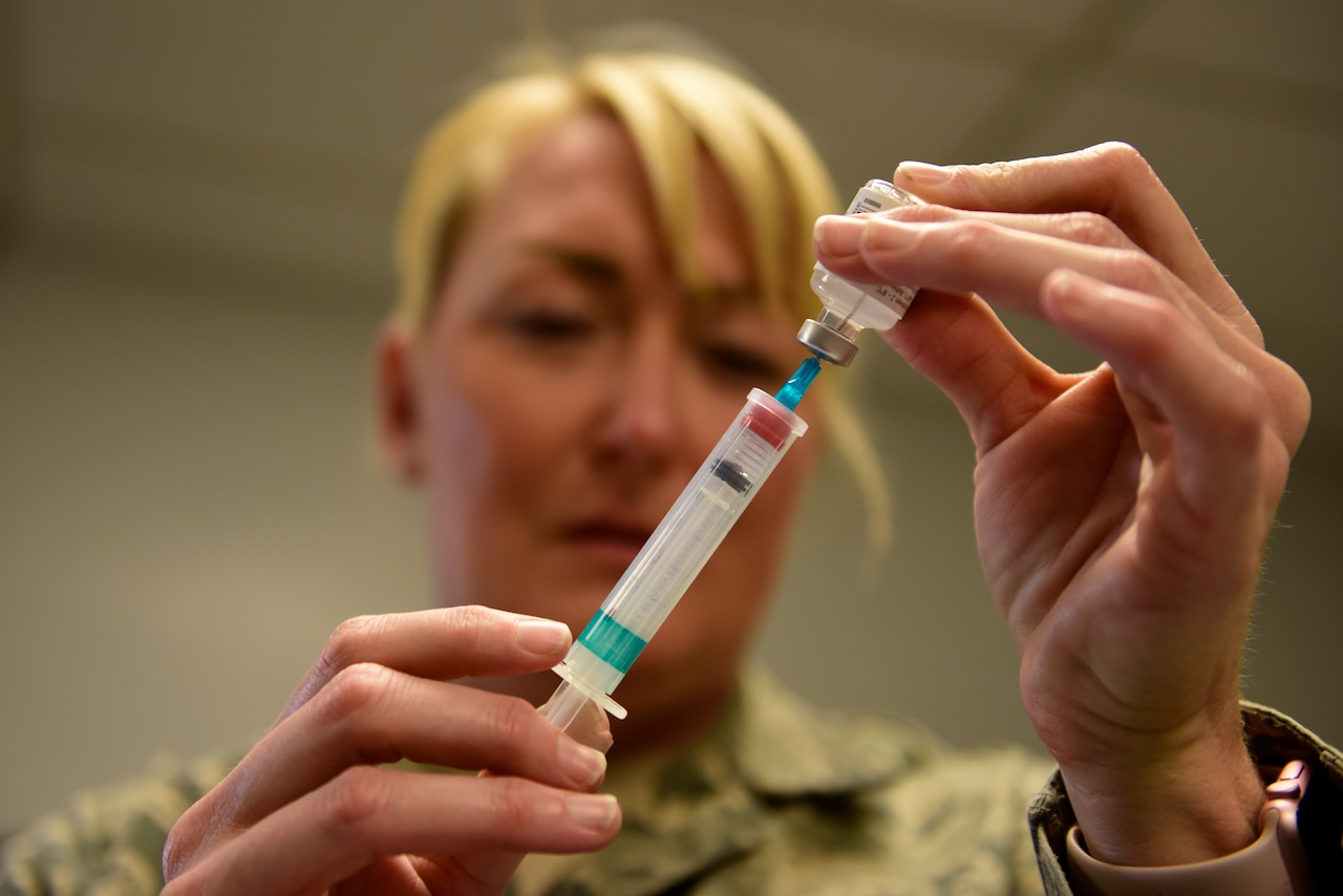 A woman in military uniform inserts a syringe into an inverted vial.
