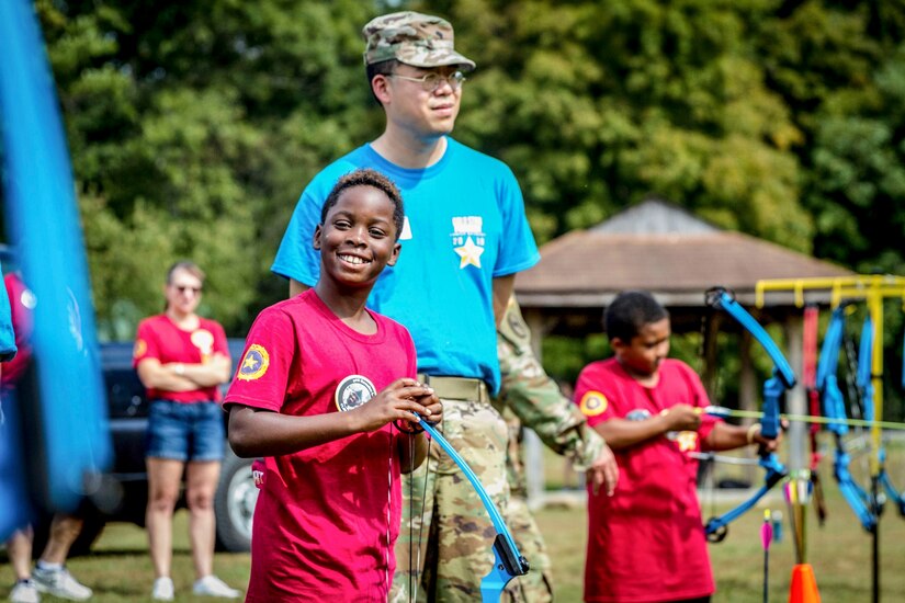 A youth holding an archery bow smiles for the camera as a soldier stands beside him on an outdoor field.