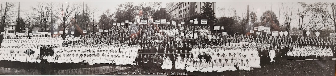A group of hundreds of people that made up the staff of the Battle Creek Sanitarium stand together outside of their building. The dress of the people is typical for workers of the early 1920s.
