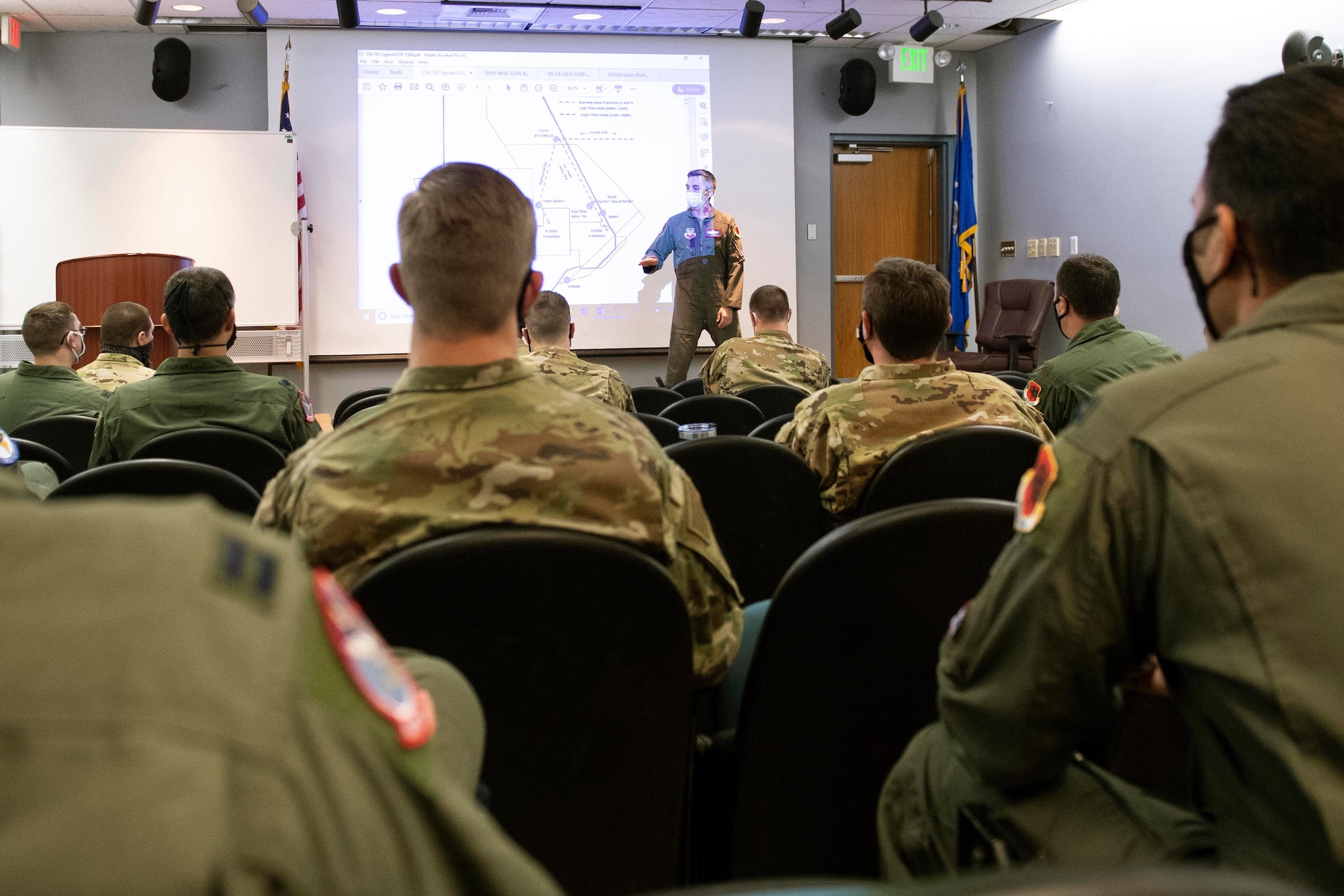 The backs of many Airmen sitting in chairs are towards the camera. The Airmen are facing an Airman presenter and a slideshow.