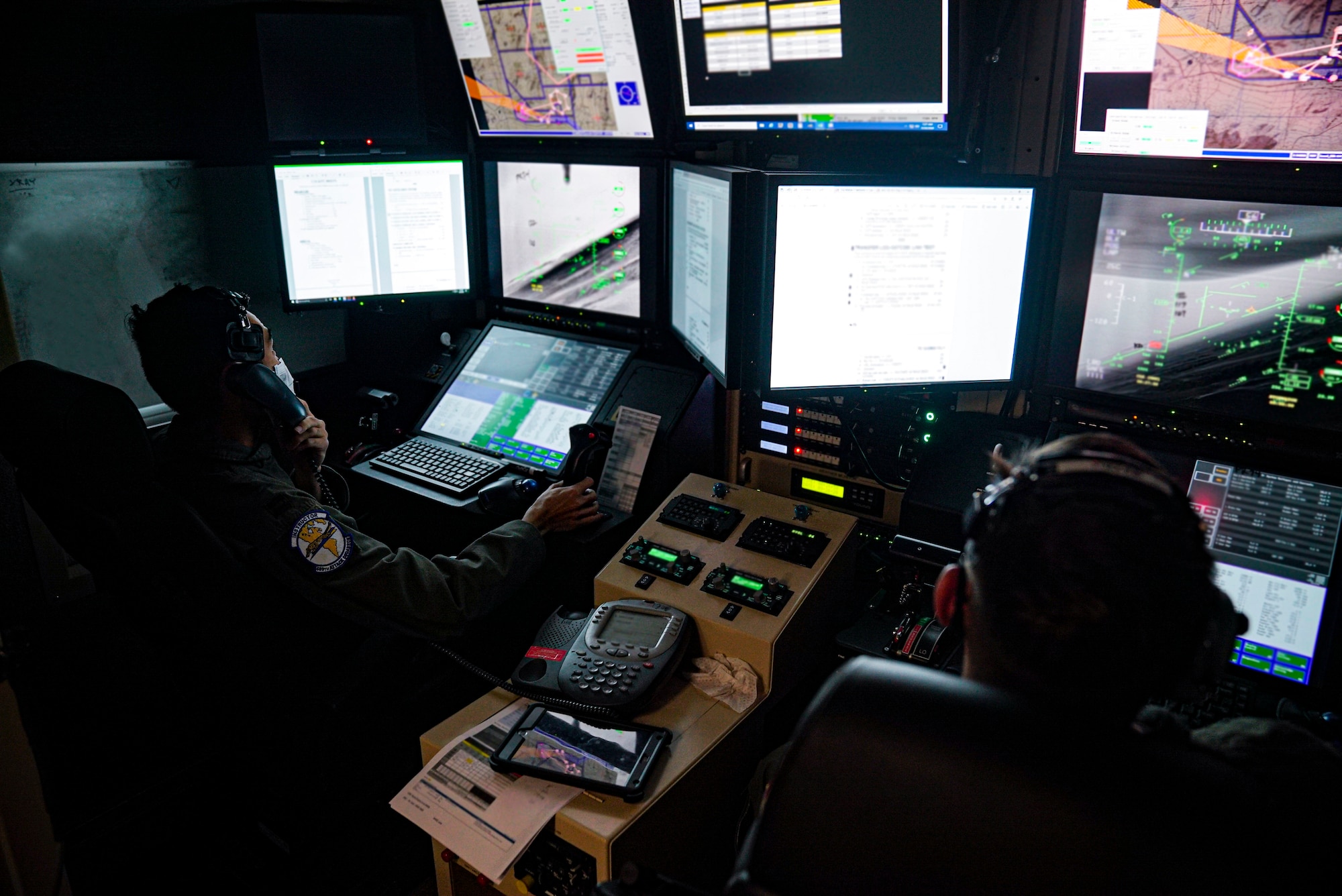 Two male Airmen in flight suits sit in an MQ-9 Reaper ground cockpit with papers and a phone between them. They are wearing a headset and are focused on the screens displaying flight information.