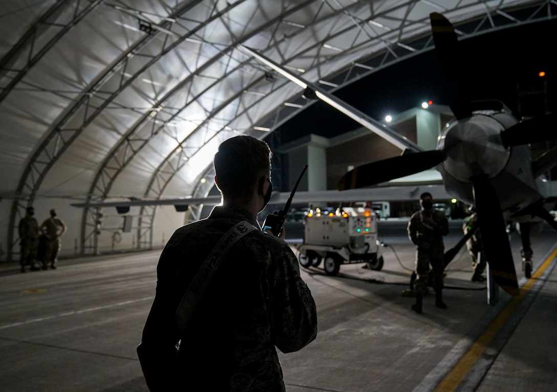 A male Airman faces the back of an MQ-9 Reaper Remotely Piloted Aircraft underneath an aircraft sunshade. The Airman is holding a walkie talkie in his right hand, and close to his face. The Airman's back is to the camera and his front is lit by a bright light. Four maintenance Airmen are working on the aircraft.
