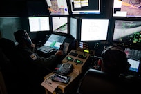 Two male Airmen in flight suits sit in an MQ-9 Reaper ground cockpit with papers and a phone between them. They are wearing a headset and are focused on the screens displaying flight information.