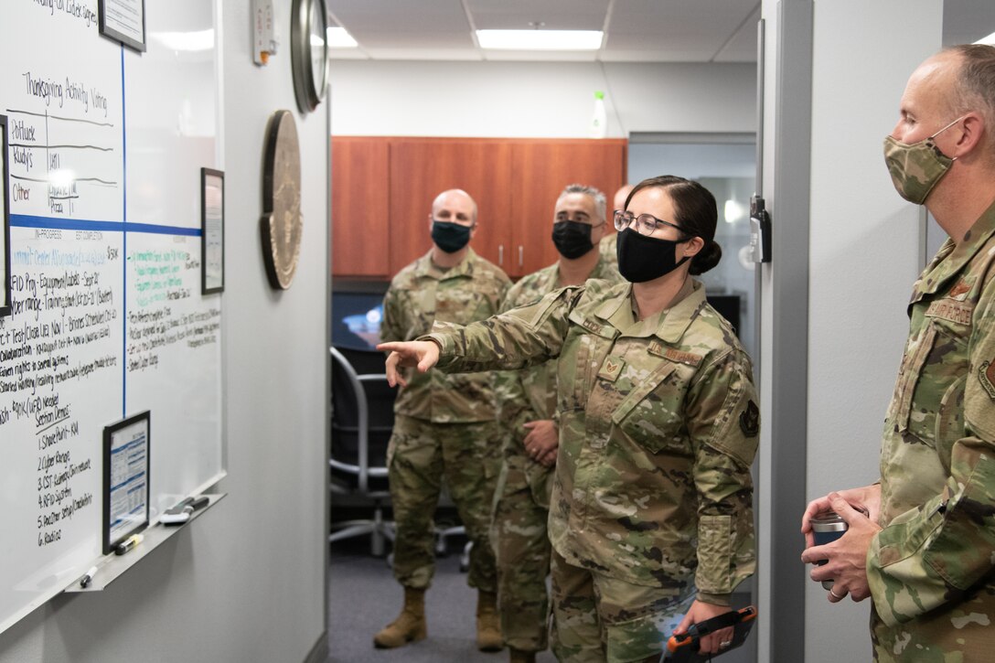 A woman gestures at a whiteboard in an office while people around her observe and listen.
