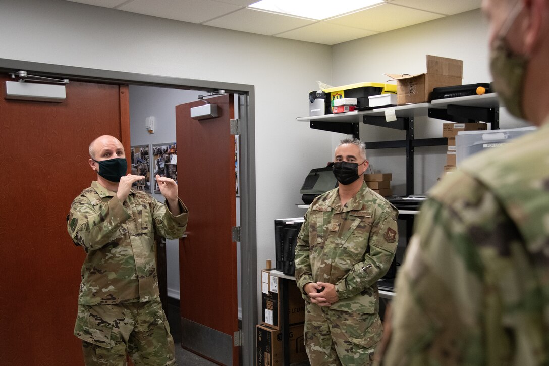 A man gestures while speaking in an equipment room while two men listen to him.