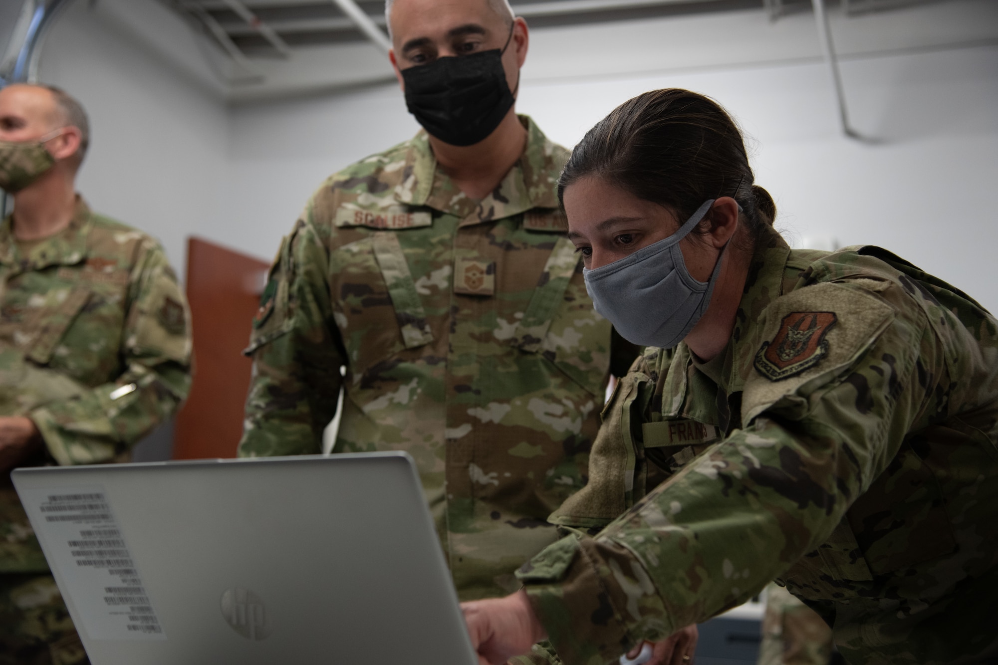 A woman wearing a blue mask points at a laptop screen while a man observes in the background.