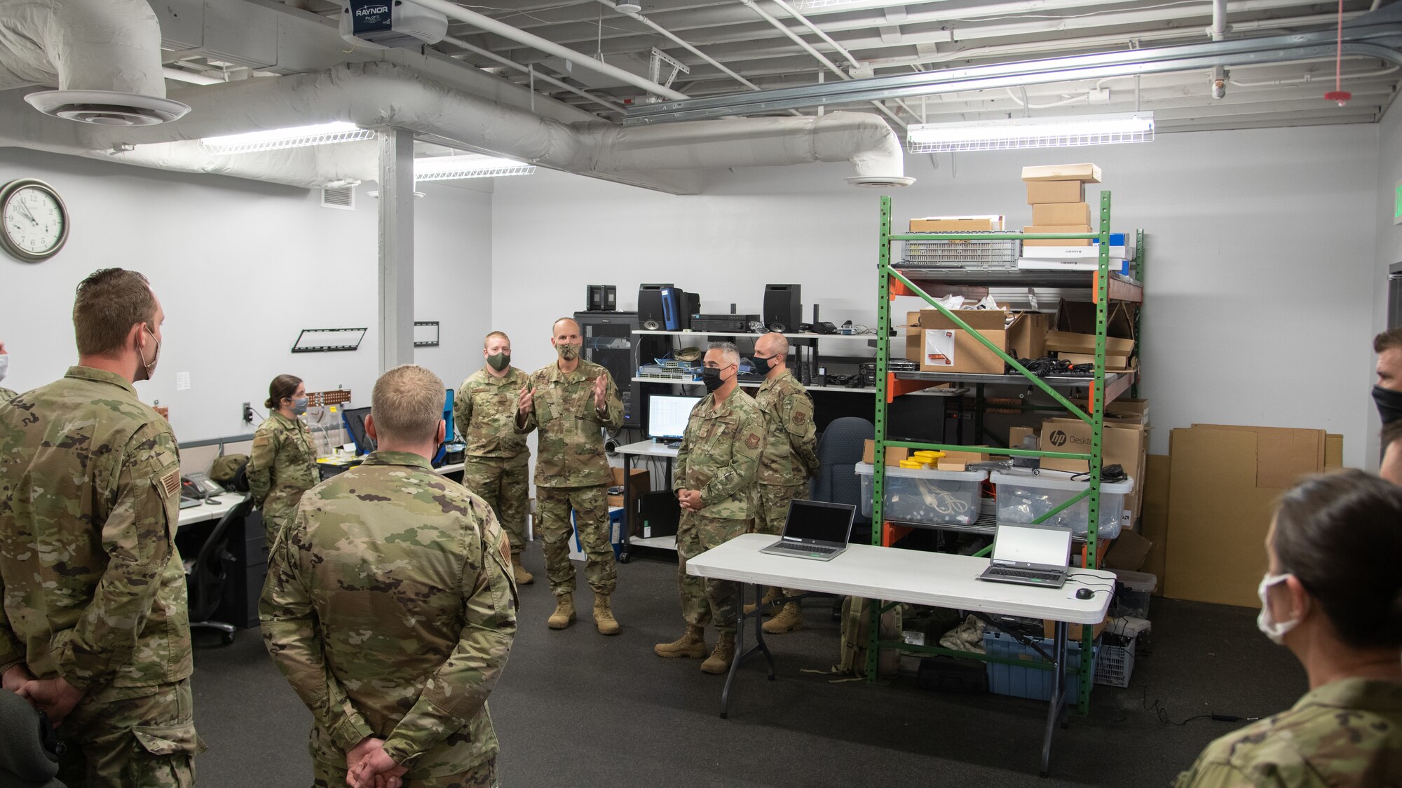 The 302nd Airlift Wing commander speaks to a group of Airmen from the 302nd Communications Flight surrounded by equipment in a room.