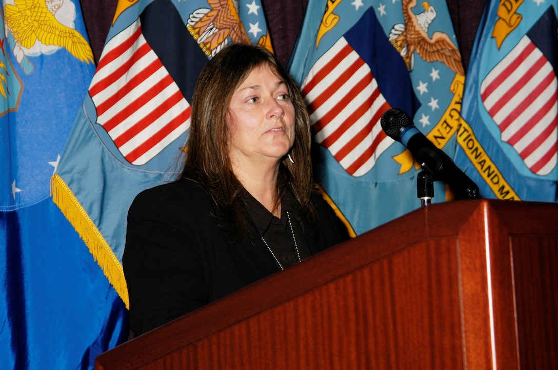 Lady in a black outfit stands behind a podium with DLA flags in the background.