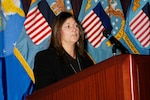 Lady in a black outfit stands behind a podium with DLA flags in the background.