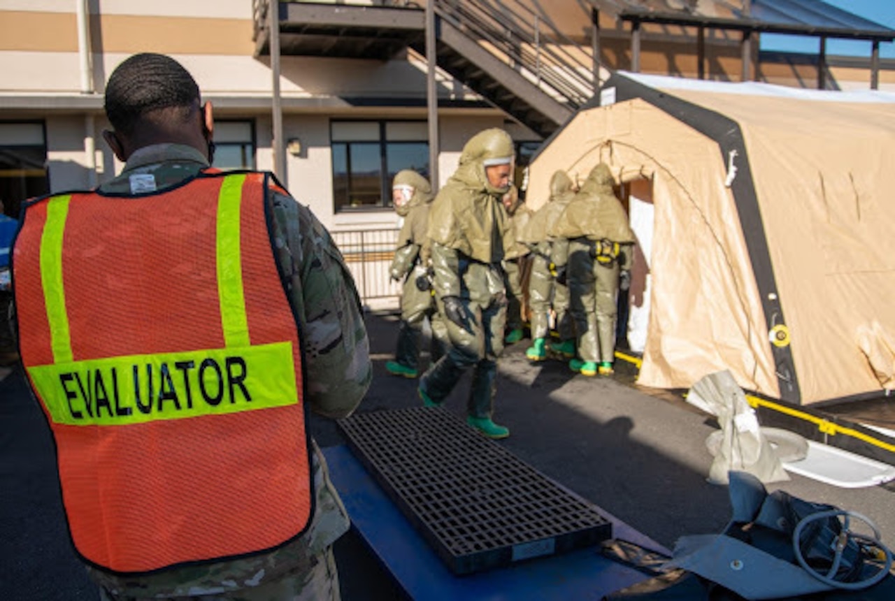 A man wearing a vest bearing the word "evaluator" watches several service members.