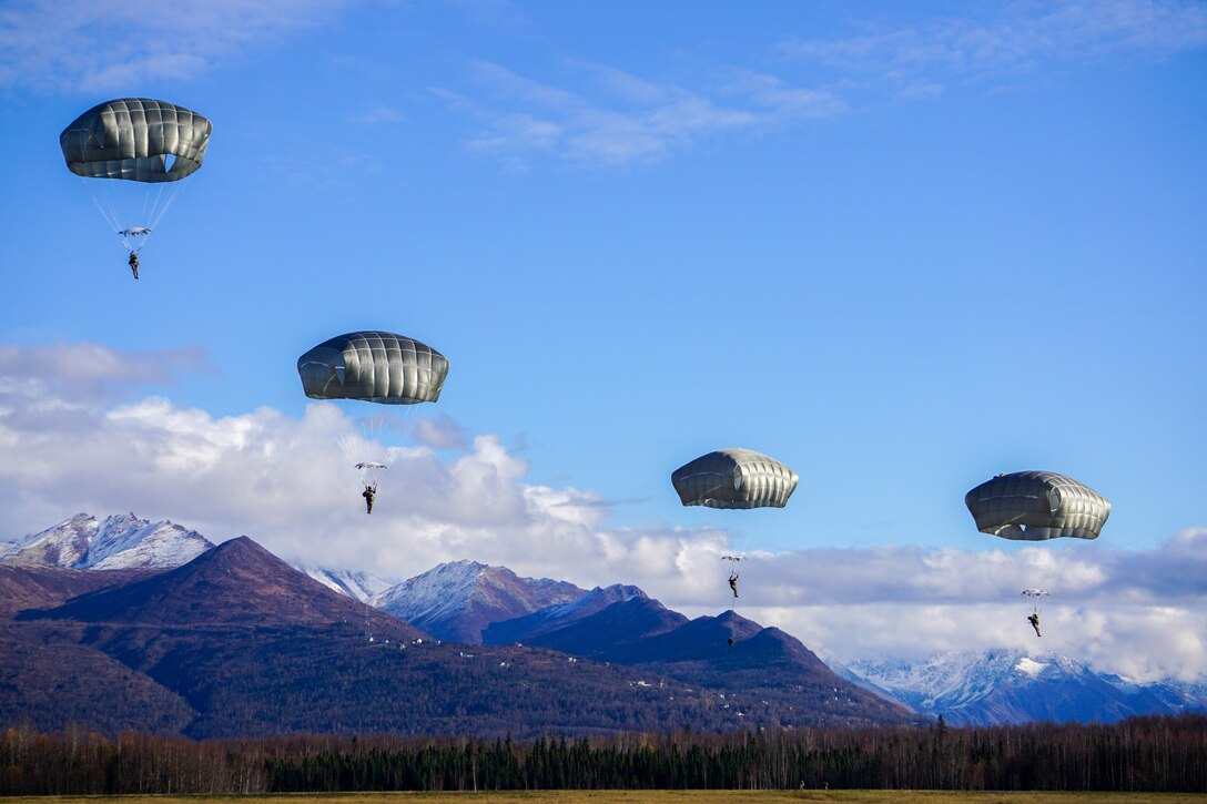 Four parachutists jump from an aircraft with mountains as a backdrop.