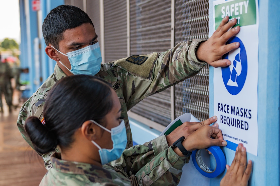 Two guardsmen -- one man and one woman -- wear face masks and tape signs requiring face masks to the outside of a building.