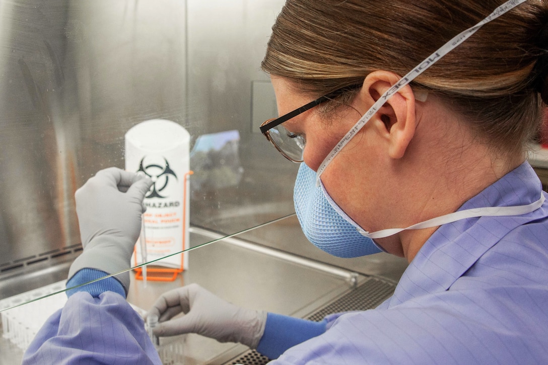 A female sailor wearing personal protective equipment holds a test kit in her right hand and a dropper in her left hand behind a glass partition.