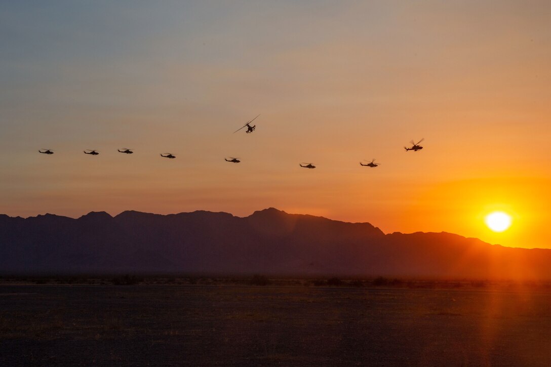 A group of helicopters fly over the mountains with the sun in the background.