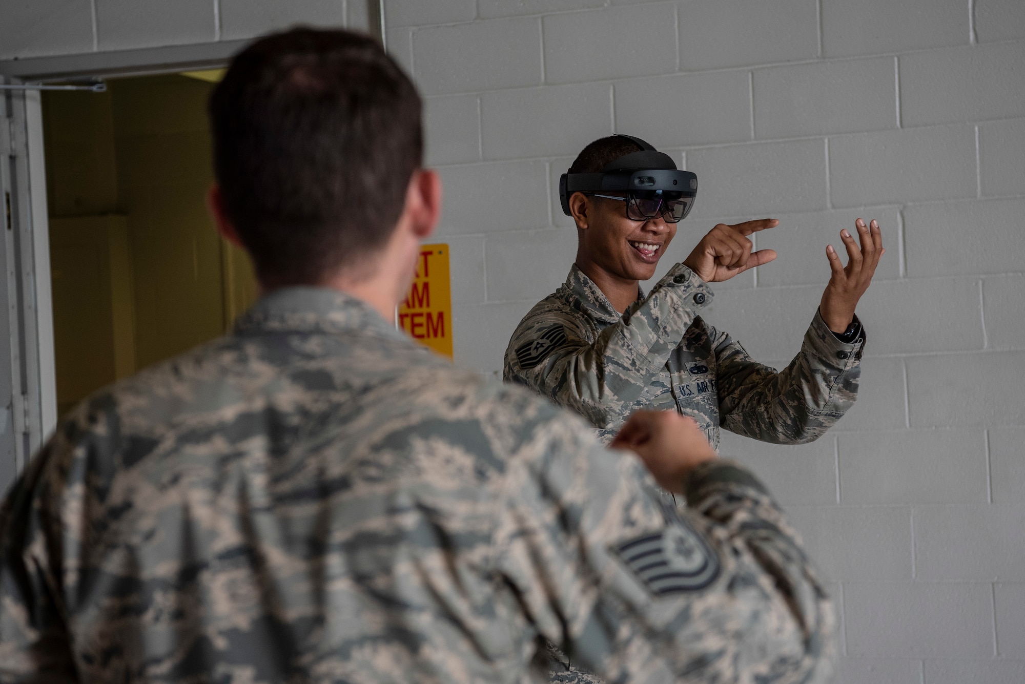 An Airman looks through an augmented reality headset at a digital technical order while another Airman directs him on how to use the gear.