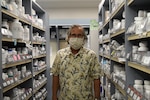 Masked pharmacist stands in pharmacy aisle.