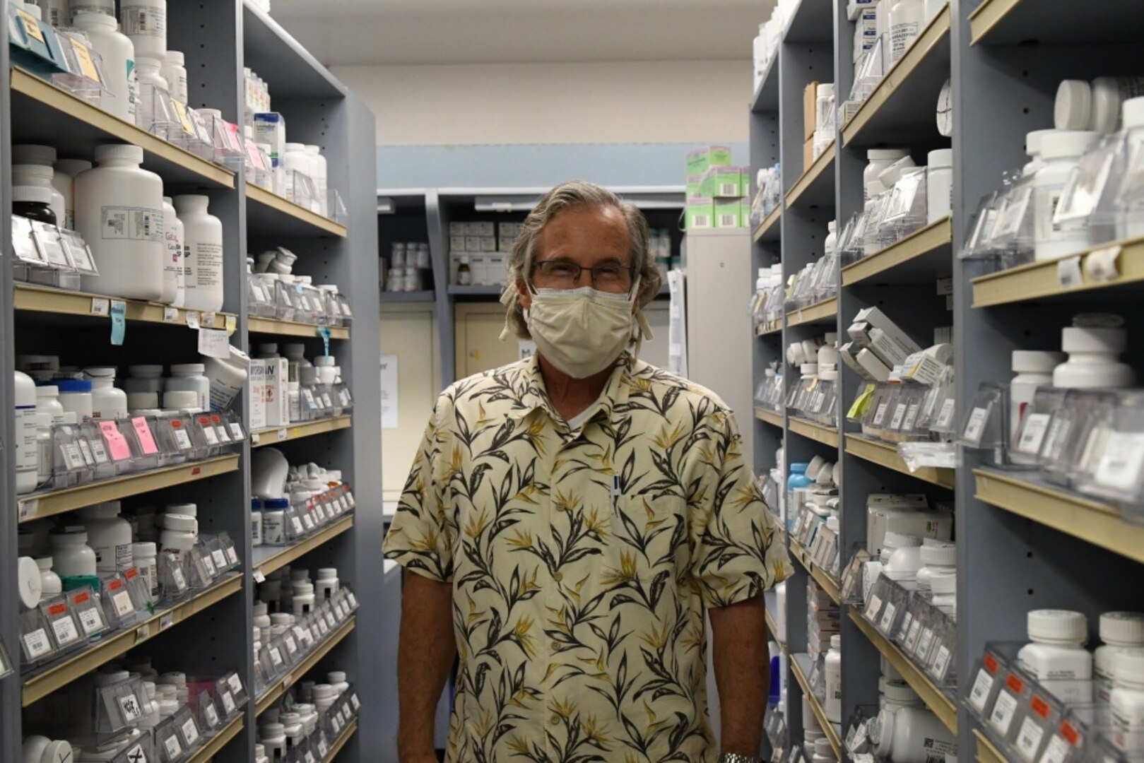 Masked pharmacist stands in pharmacy aisle.