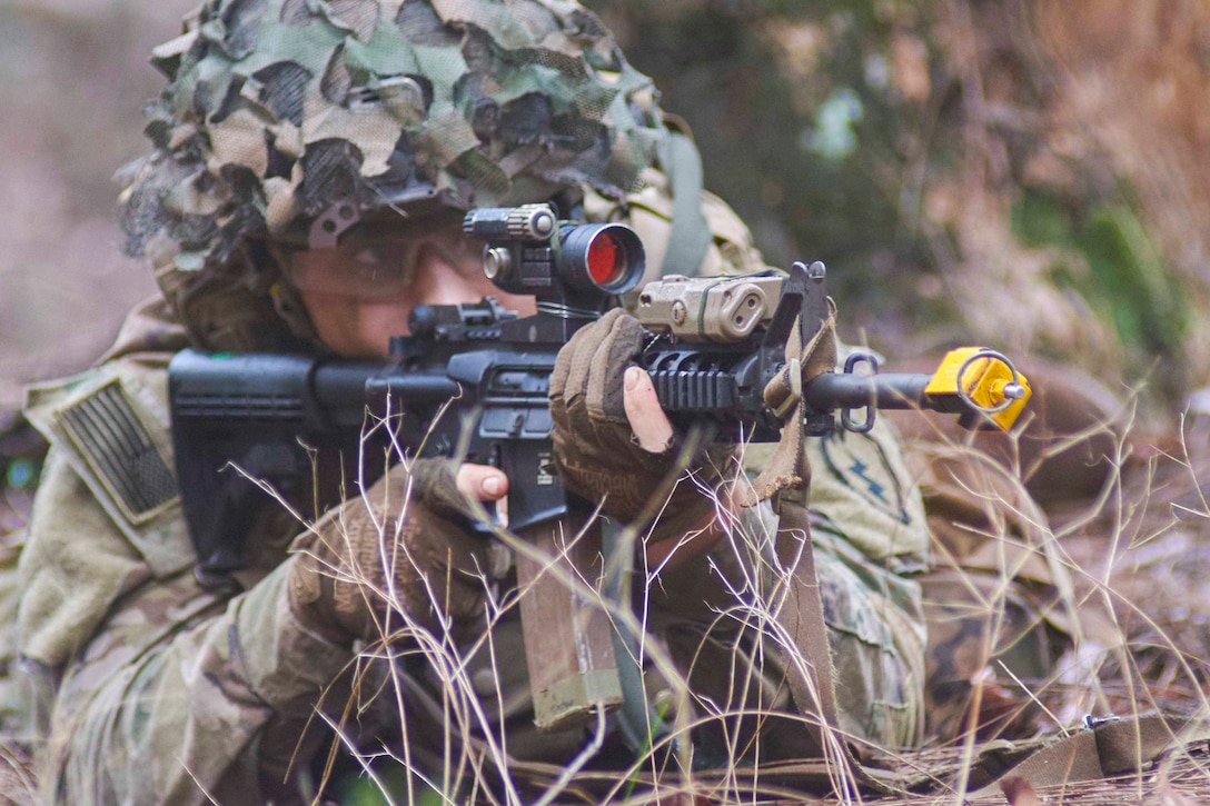 A soldier looks through the scope of his weapon while camouflaged in dry brush.