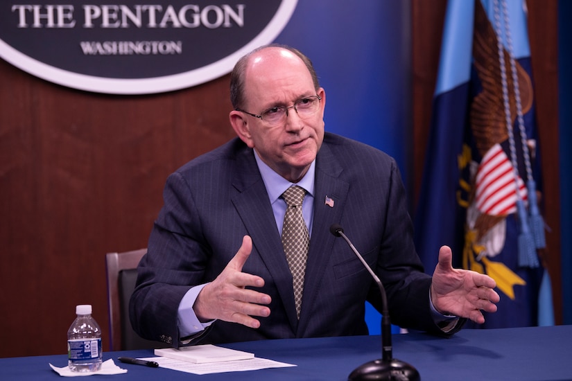 A man sits behind a table.  Behind him, a sign reads, "The Pentagon - Washington."