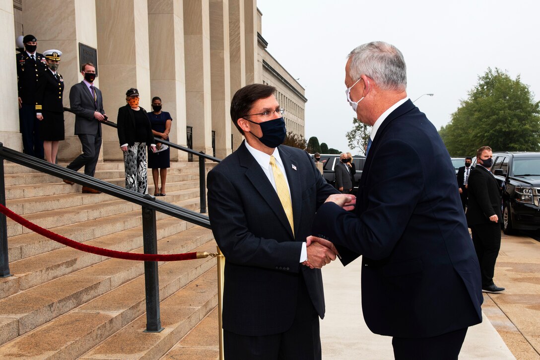 Two men dressed in suits and wearing face masks greet one another with a handshake outside a large building.