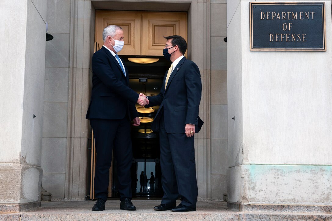 Two men dressed in suits and wearing face masks greet one another with a handshake outside a large building.