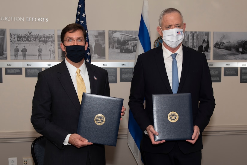 Two men dressed in suits and wearing face masks stand next to one another; each holds a binder with a seal in the middle.