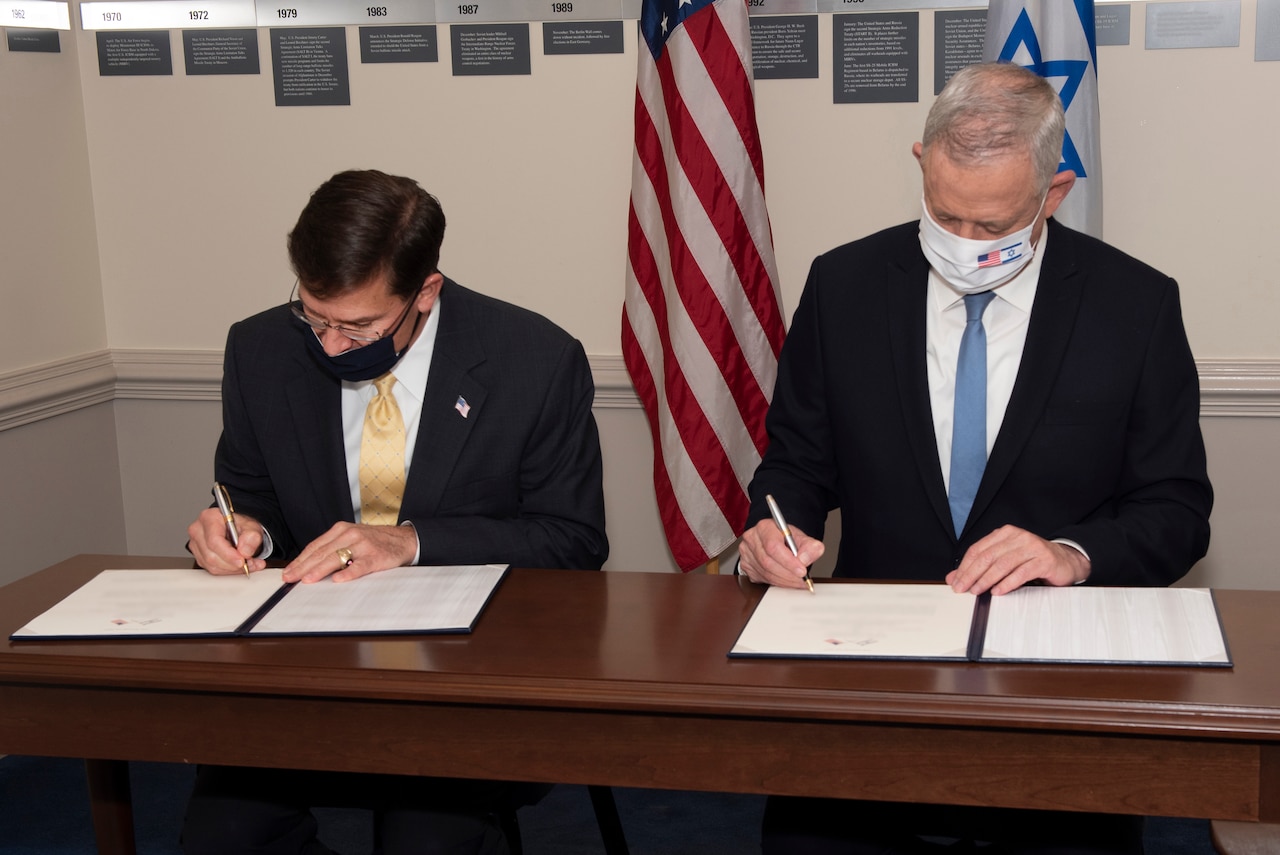 Two men dressed in suits and wearing face masks sit next to one another; each is signing a document.