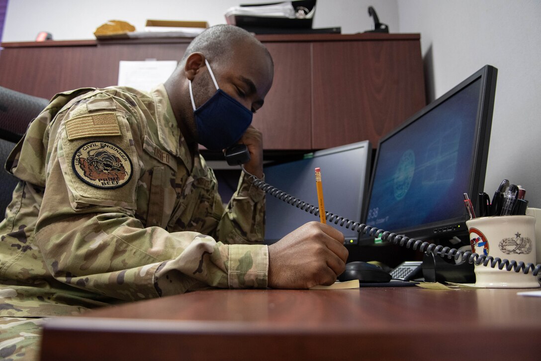 An Airman sits a desk, speaking on the phone and taking notes.