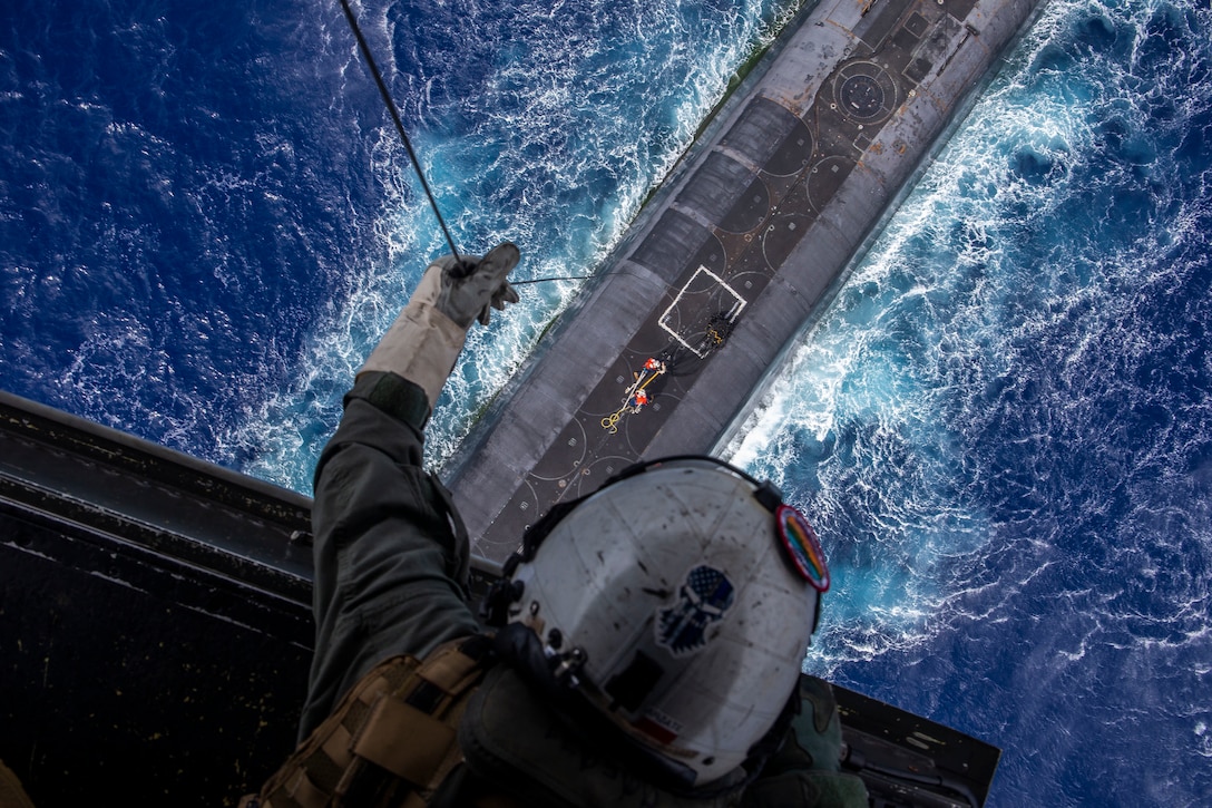 A Marine, shown from above, lowers a line from an open aircraft to a submarine in water.