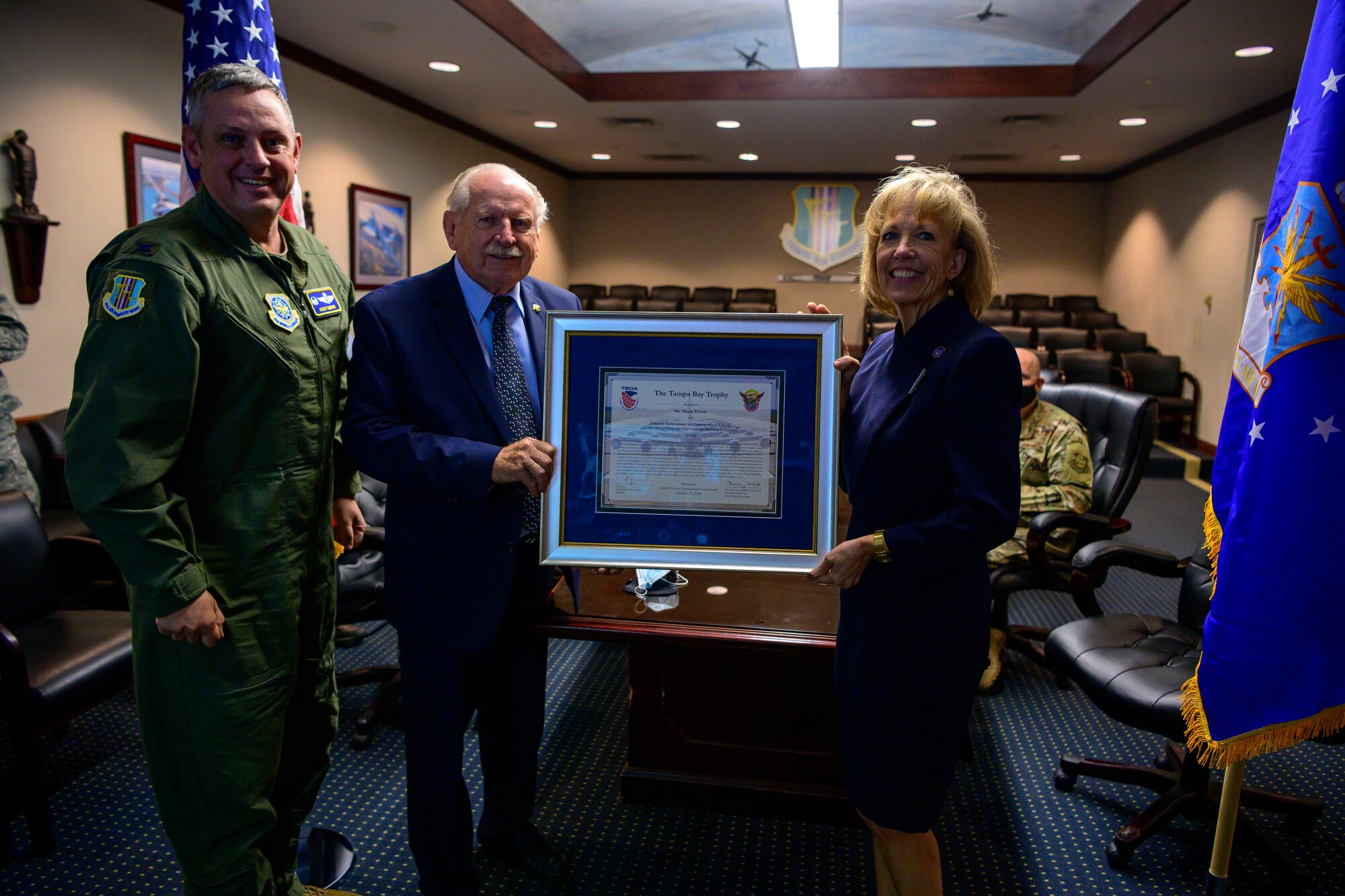 A woman in a navy dress holds a large framed certificate being presented to her by two men.