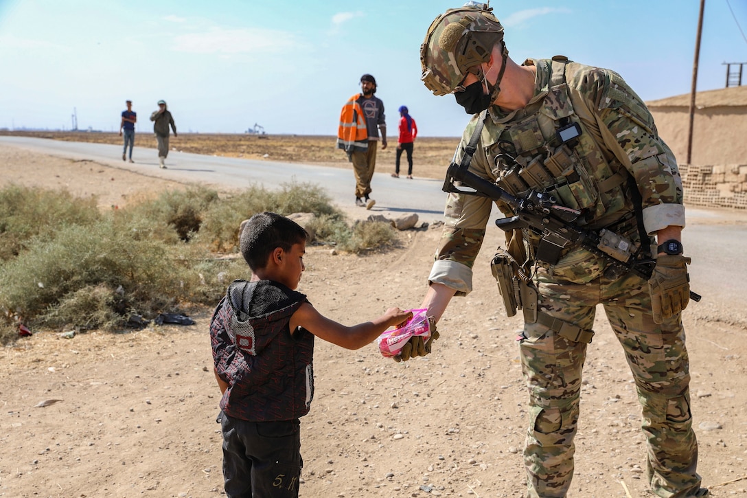 A soldier hands candy to a child in desert-like area as others walk nearby.