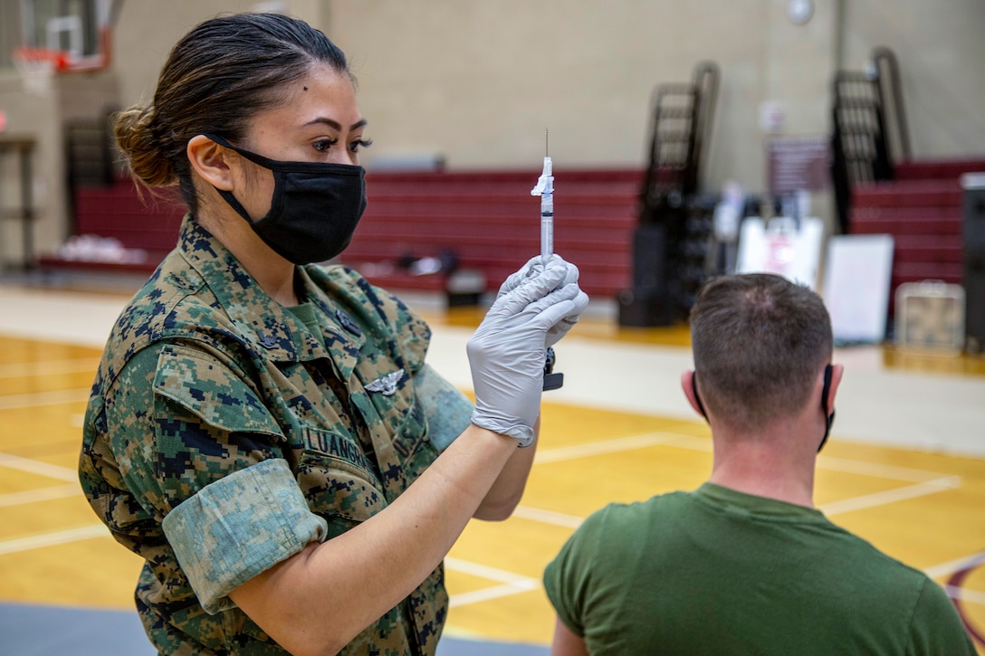 A Marine wearing a mask stands and holds up a syringe as a Marine sits in a chair beside her.