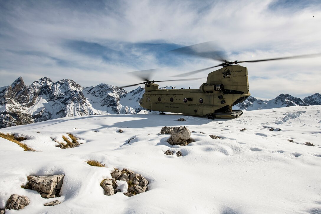 An Army helicopter sits on a snowy mountain field overlooking other snow-capped peaks.
