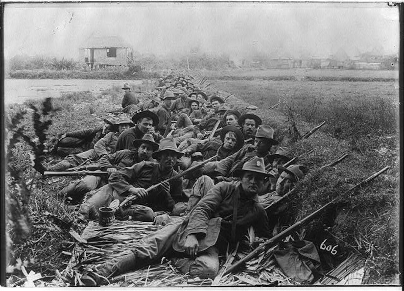 Dozens of soldiers with guns lay in a trench.