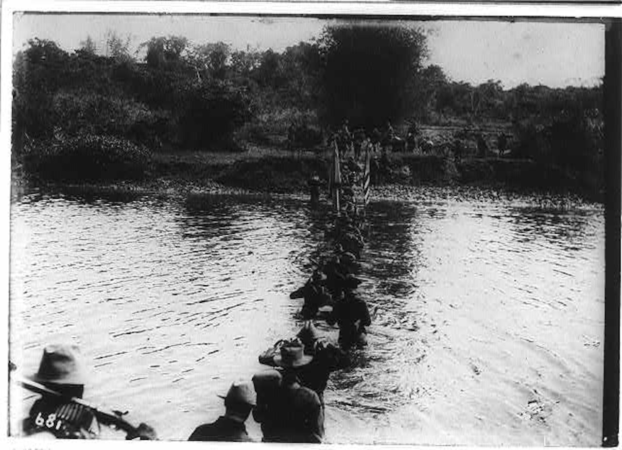 A row of men cross a waist-high river.