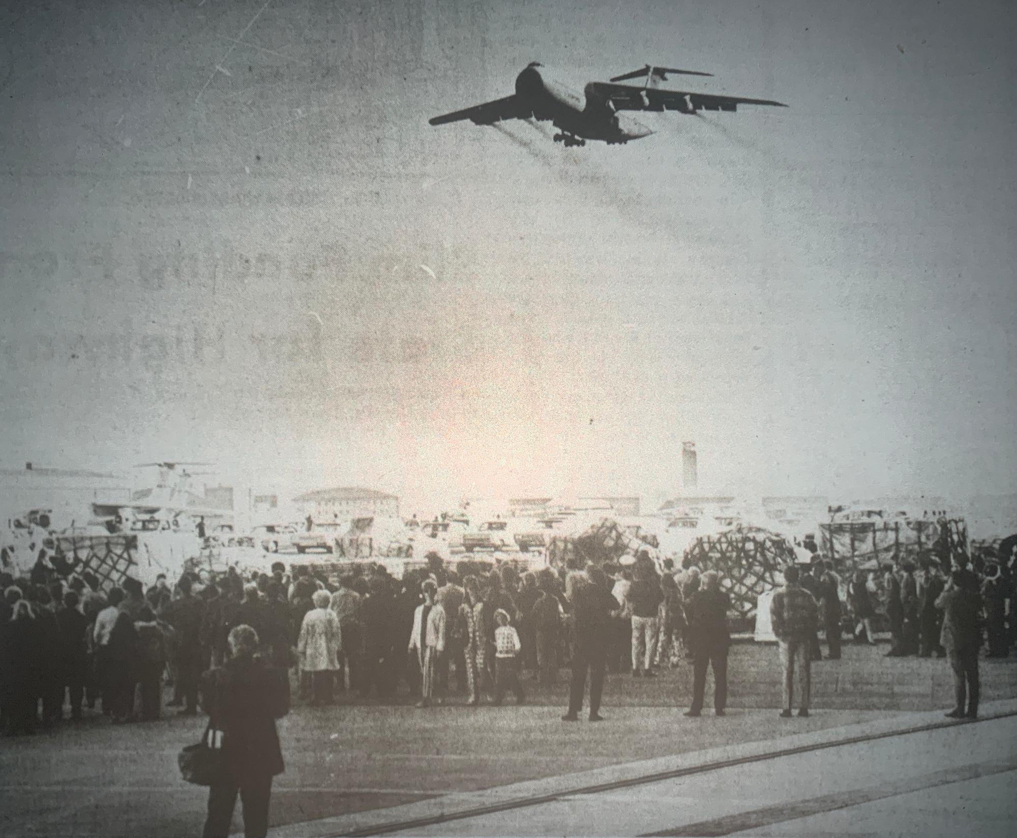A very old image with a crowd in front and in the top of the image you can see a C-5 with its landing gears open about to land on the flight line.