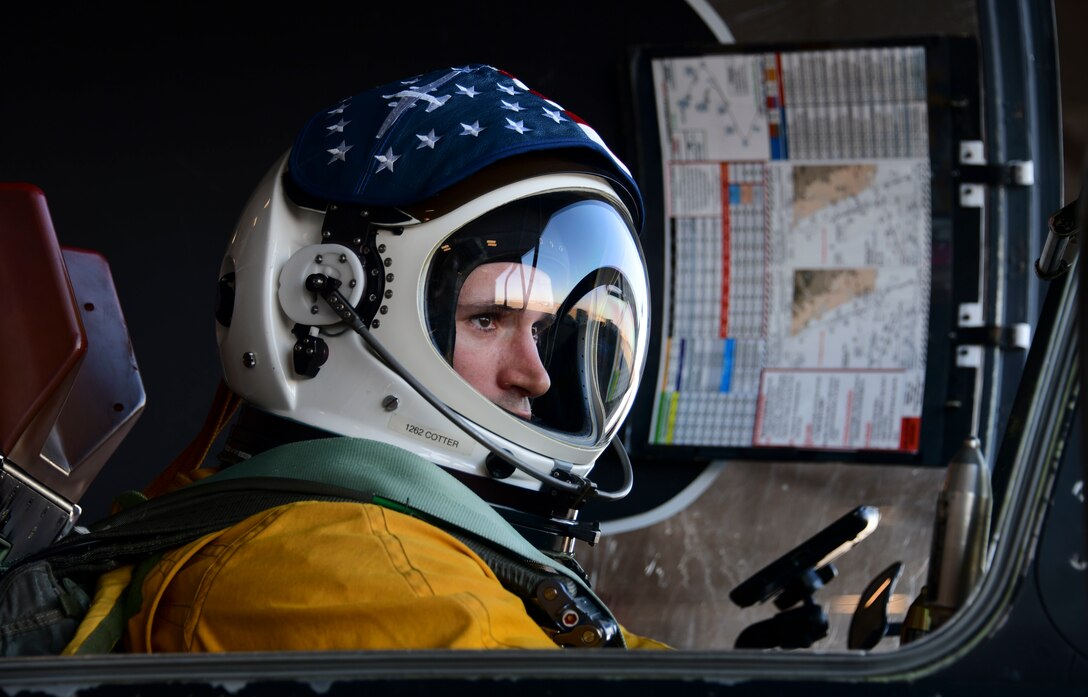 An Air Force pilot in a yellow flight suit and large white helmet sits in a cockpit.