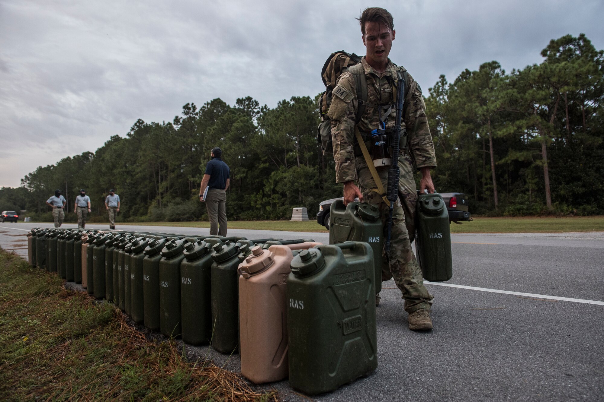 A lone Special Tactics tactical air control party candidate carries a pair of jerry cans past a long row of jerry cans during an assessment