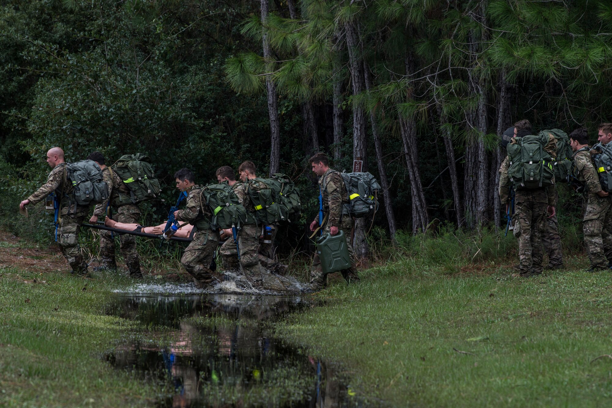 Four Special Tactics tactical air control party candidates carry a litter across a creek in the distance as another candidate crosses the picture closer to the camera. They appear to be moving quickly.