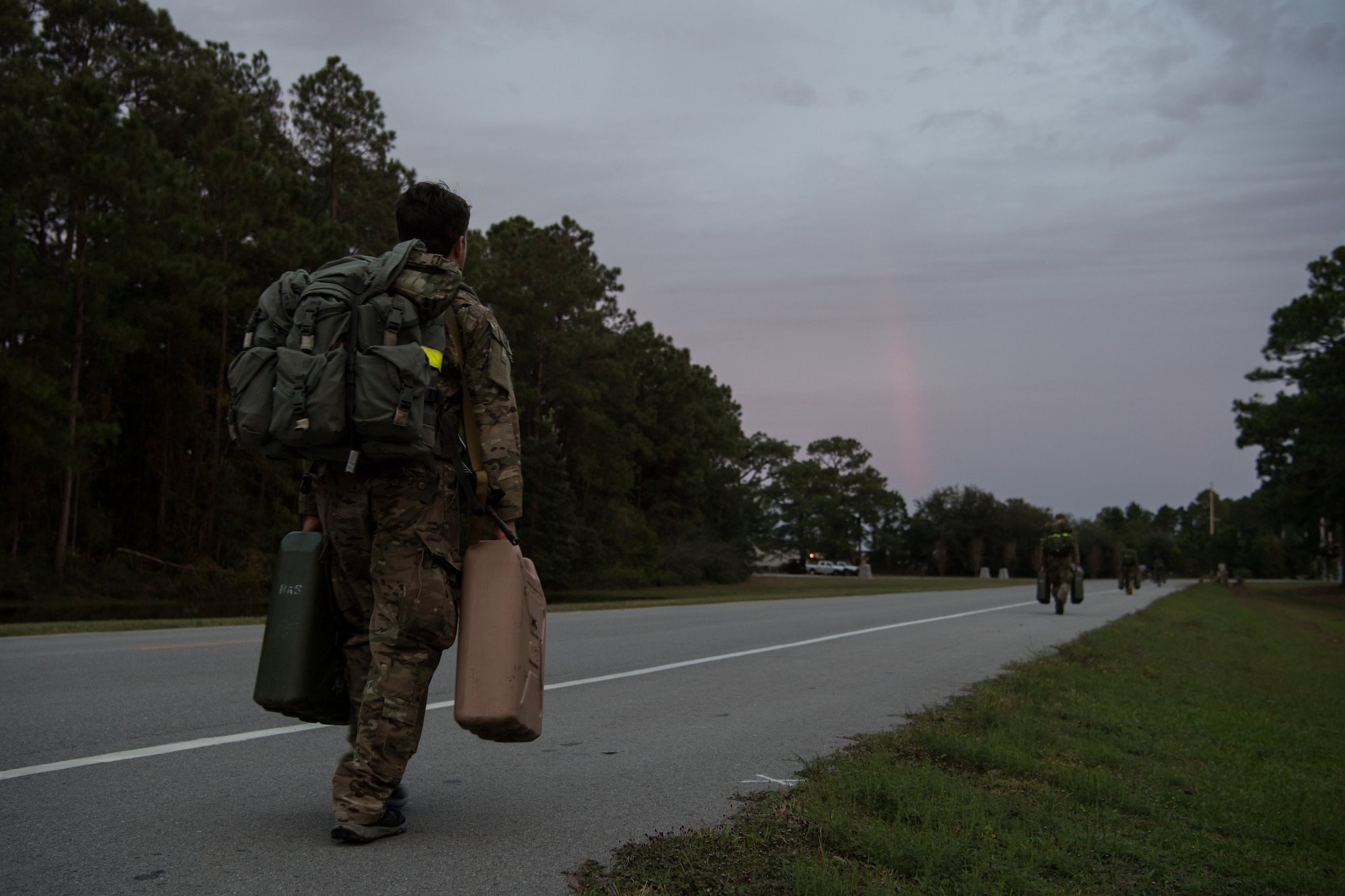 A lone Special Tactics tactical air control party candidate walks beside the road carrying a pair of jerry cans as part of his selection assessment. Other candidates carrying pairs of jerry cans can be seen much further down the road.