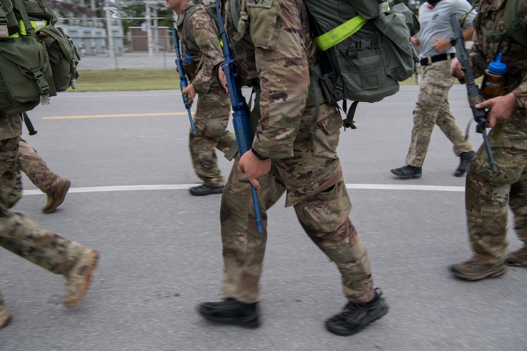 Special Tactics tactical air control party candidates carry rifles and heavy packs while quickly walking beside a road.