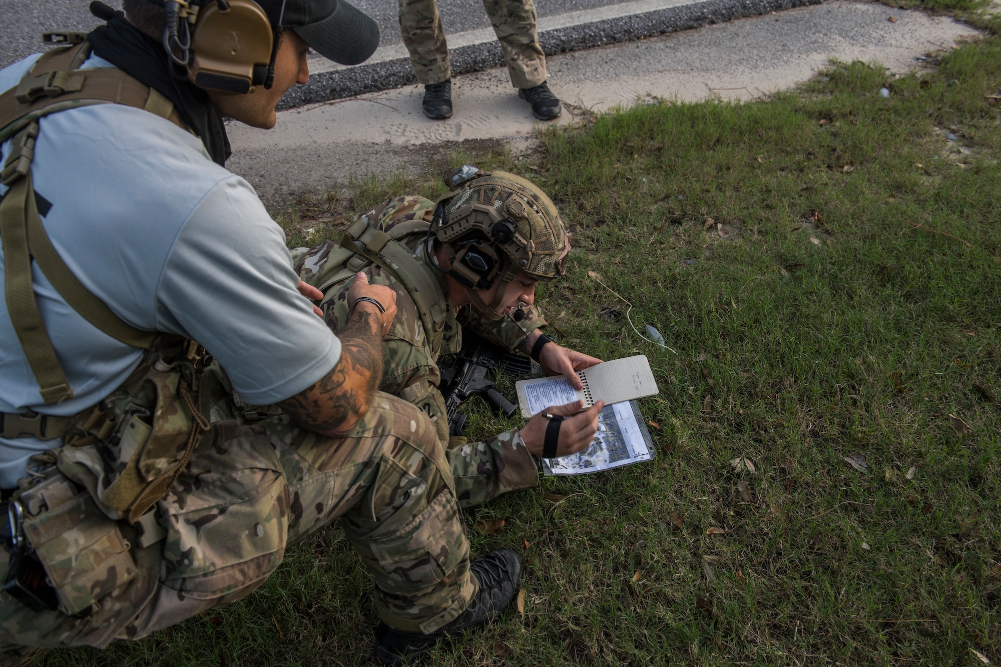 A pair of tactical air control party candidates participate in a call for fire scenario. One crouches on the ground looking at a map while a light-blue shirted cadre member kneels beside him and talks on his headset.