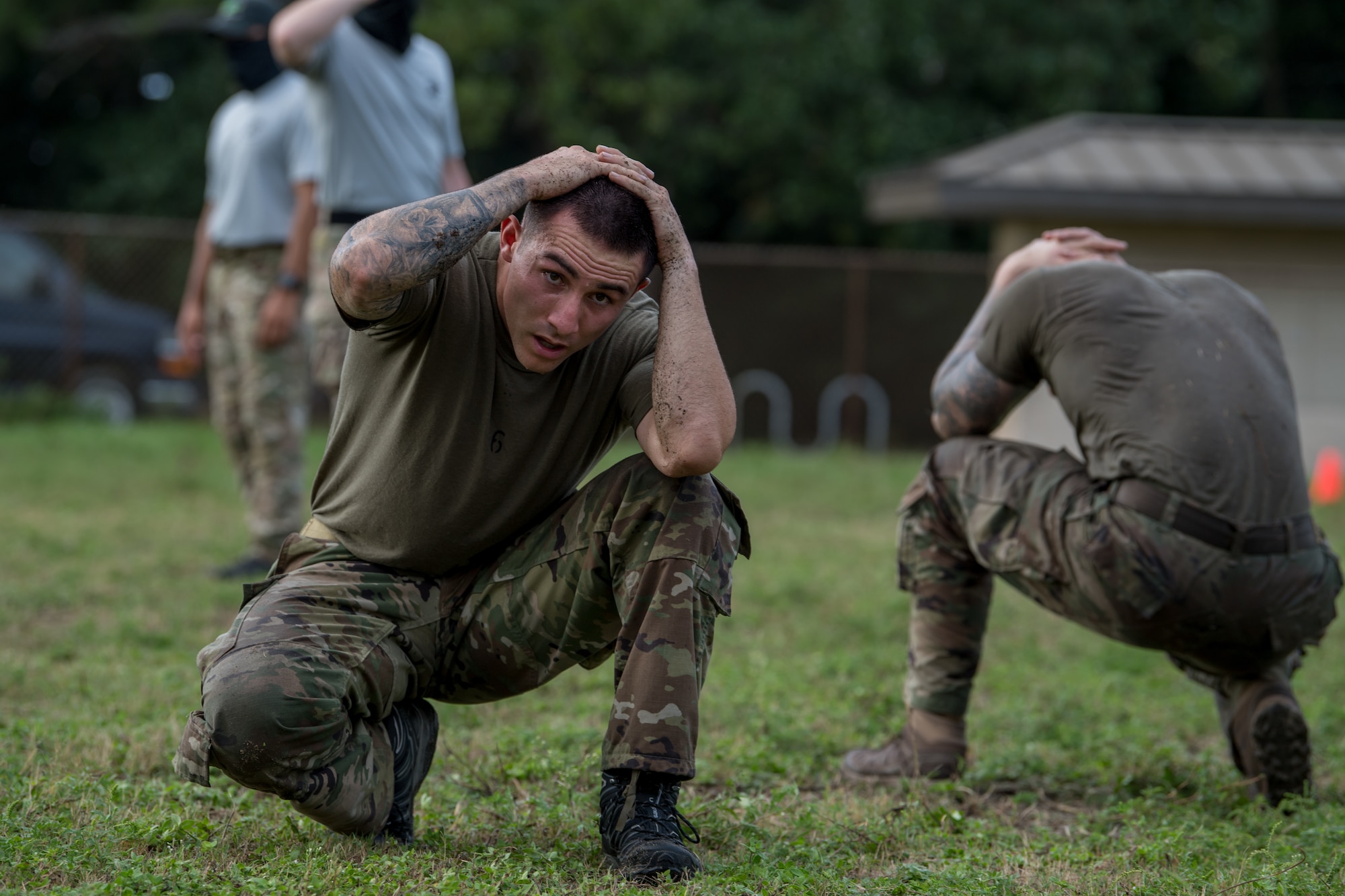 A Special Tactics tactical air control party candidate conducts an exercise walking along the grass in a crouched position with his hands behind his head.
