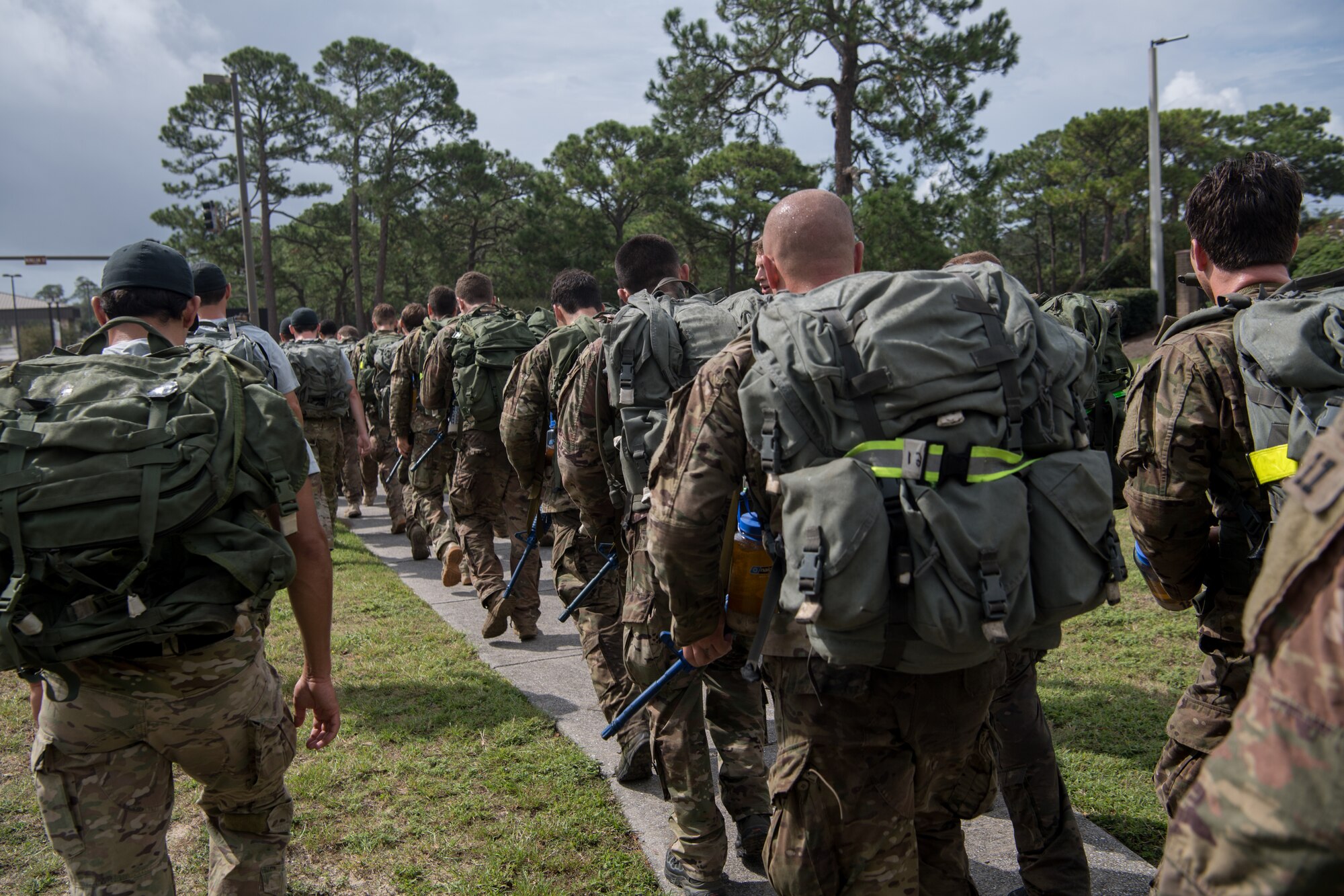 Special Tactics tactical air control party candidates and cadre walk along side-by-side carrying heavy ruck sacks, forming several lines stretching into the distance.