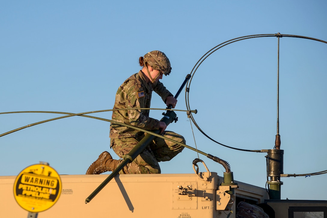 A soldier kneels on top of a vehicle and adjusts radio equipment.